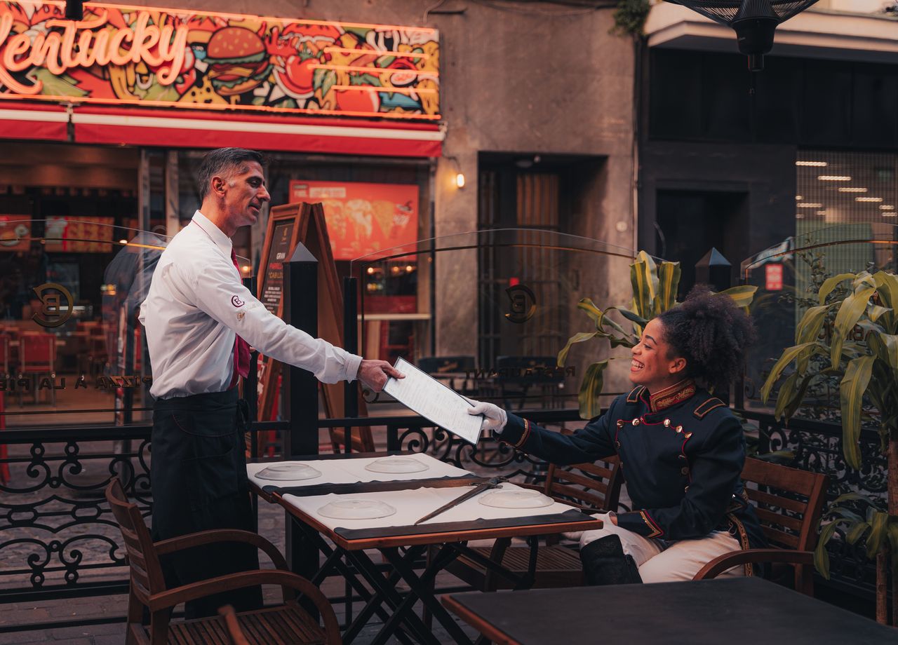 Services A waiter serves a menu to a seated customer at an outdoor restaurant with colorful signage.